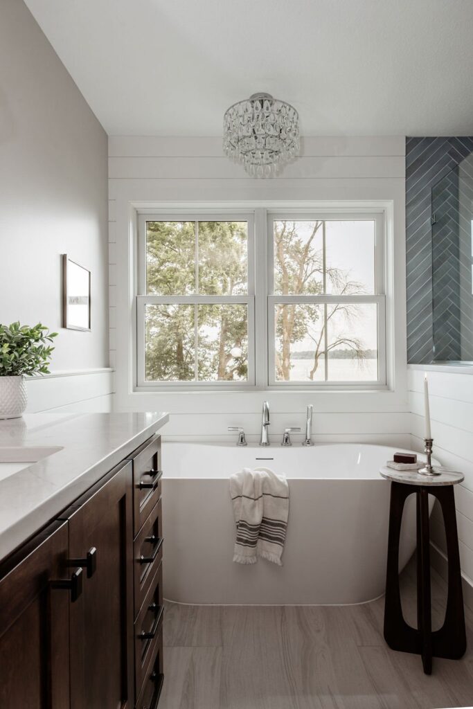 Bathroom with a freestanding white soaking tub under a shiplap wall and a double window overlooking trees, a dark wood vanity with black hardware, a crystal flush-mount ceiling light, and a small round side table.