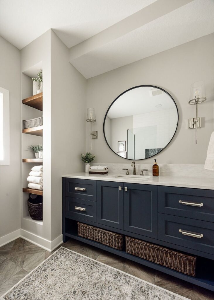 Bathroom with navy blue shaker-style vanity cabinets, a round black-framed mirror, white quartz countertop, and a built-in wood shelving alcove holding folded towels and woven baskets, over a patterned area rug.