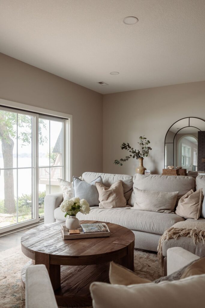 Finished living room after an interior design install, with a light gray sectional sofa styled with cream and beige throw pillows, a round wood coffee table topped with hydrangeas, a sliding glass door showing a lake view, and an arched mirror on the far wall.