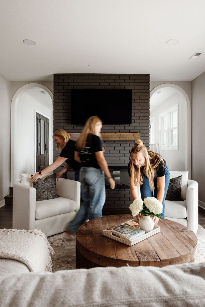 Interior designers styling a living room on install day, arranging pillows and flowers on the coffee table in front of the fireplace.