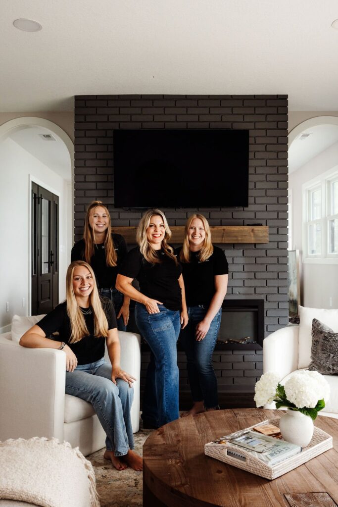 Interior design team posing together after finishing a living room install, in front of a dark charcoal brick fireplace with a mounted TV, with white hydrangeas styled on the coffee table.