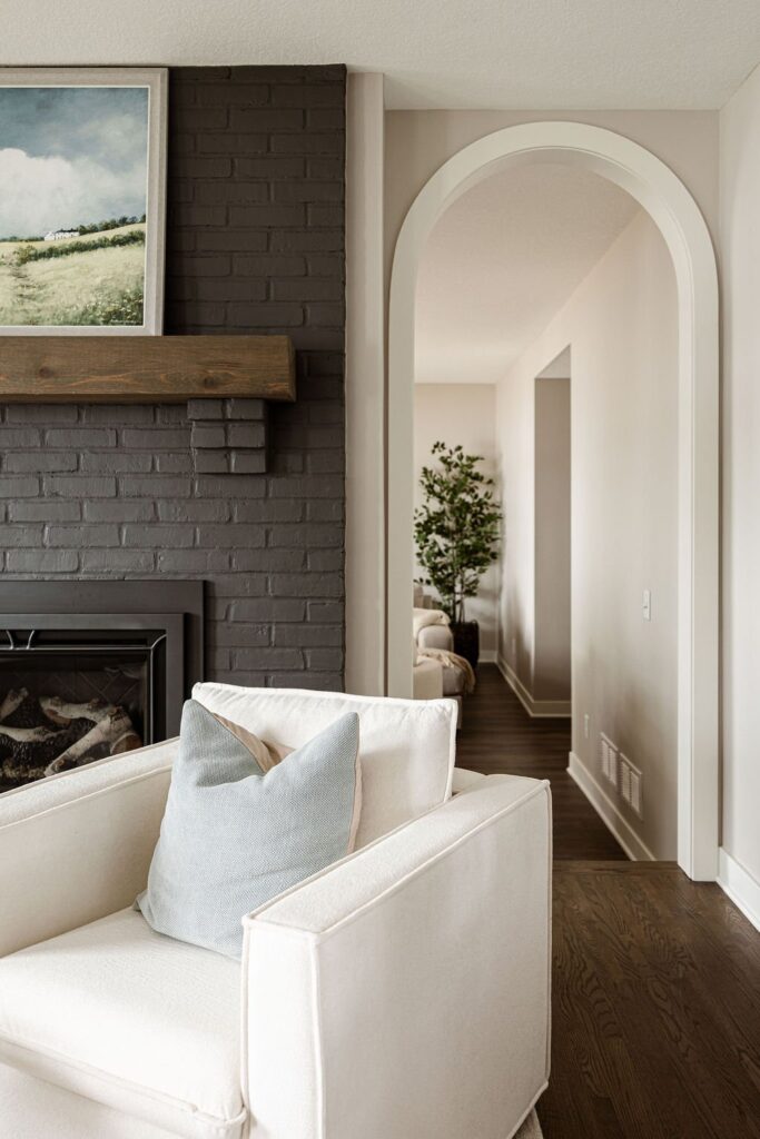 Living room corner with a dark charcoal painted brick fireplace, wood mantel, framed landscape painting, and a white swivel armchair with a sage green throw pillow, next to an arched doorway opening to a hallway with a potted tree.