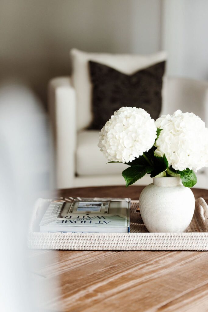 Close-up of a round wood coffee table styled with a woven tray holding a white textured vase of hydrangeas and a stack of home decor books, a white armchair with a black patterned pillow blurred in the background.